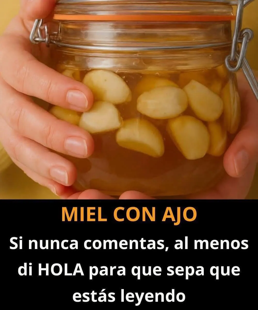 Frasco de vidrio con dientes de ajo cubiertos con miel natural sobre una mesa de madera, acompañado de una cuchara y una cabeza de ajo fresco.